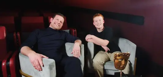 A man with short cropped dark hair and wearing all black sat in a large grey tub chair talking to a young man with ginger hair and wearing a black jumper.