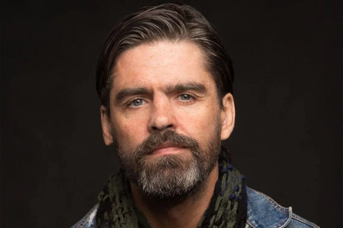 A white man with thick dark hair and a full beard with grey in it. He is wearing a denim top and a black scarf.