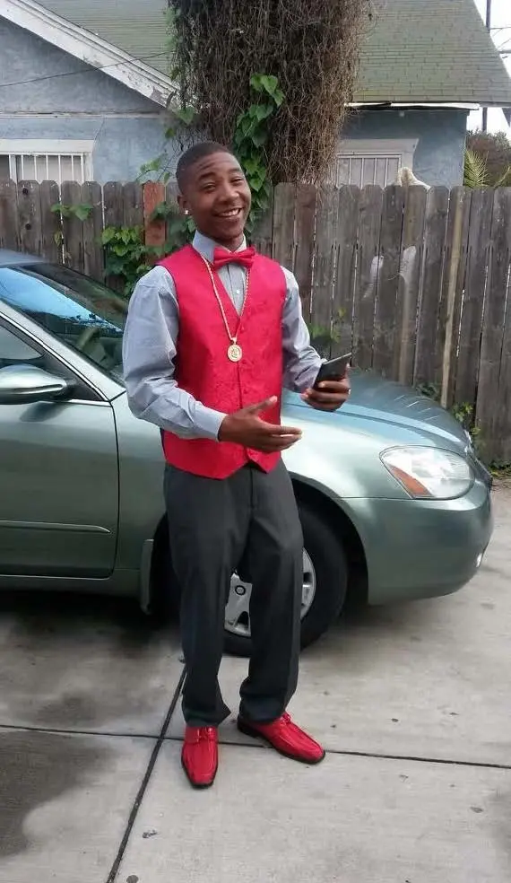 A young black man in a blue shirt, smart trousers and a red waistcoat and bow tie. He is stood in front of a silver car.