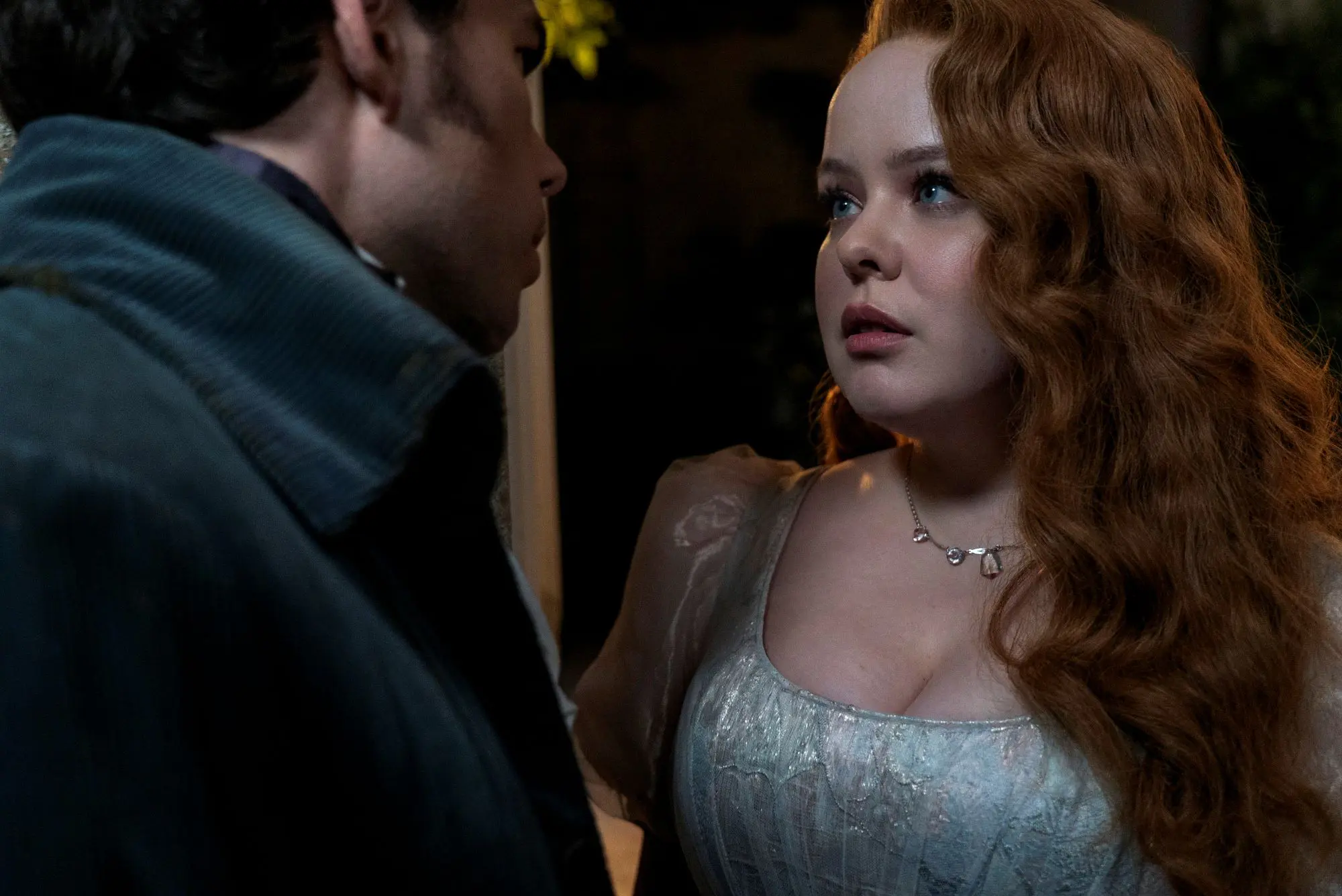A man and a woman in regency era clothing standing with their faces really close together. The man is white with thick dark hair wearing a blue velvet jacket and the woman is white with long red wavy hair wearing a silvery bodice dress.