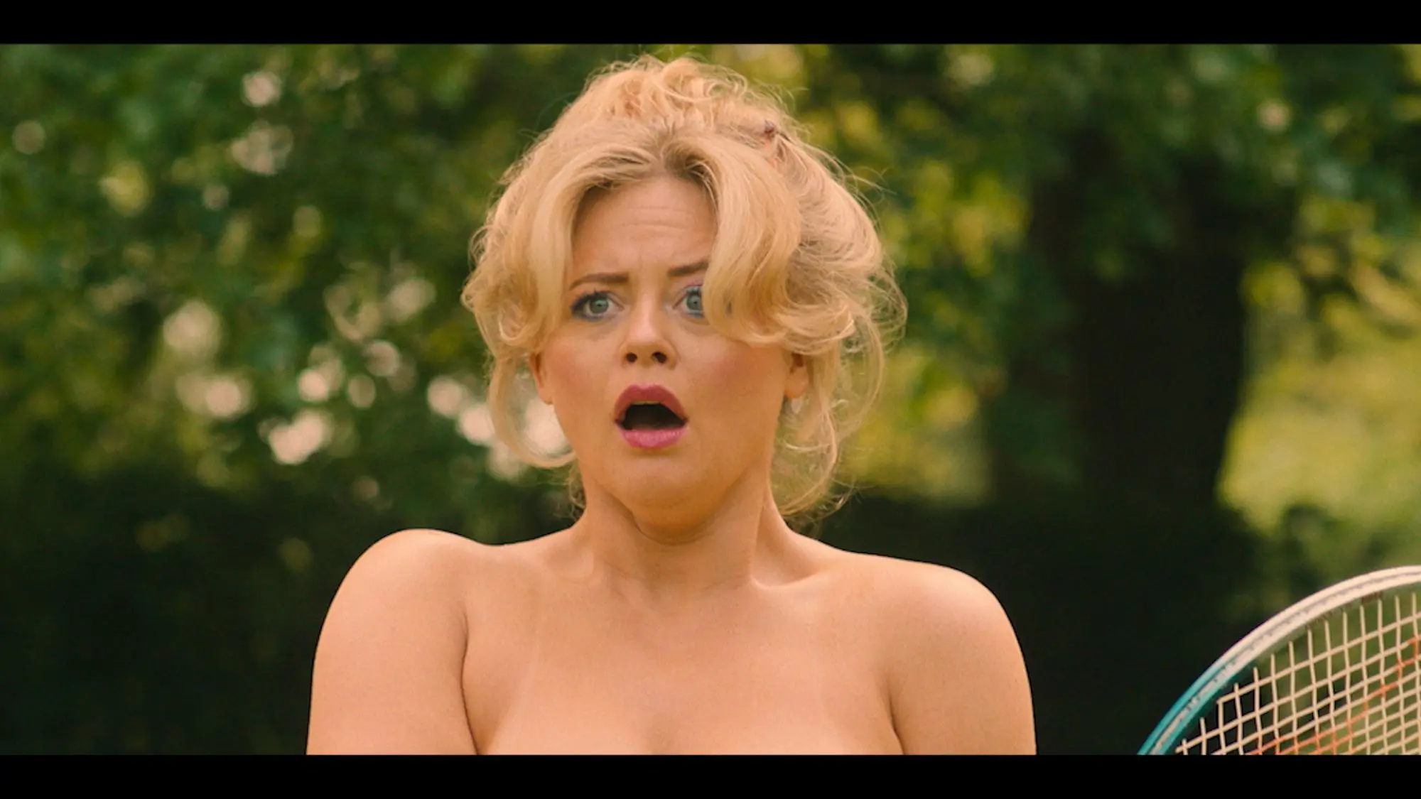 A young blonde woman with her hair tied in a messy bun and a grown out fringe. She appears to be stood outside and looks like she is topless.