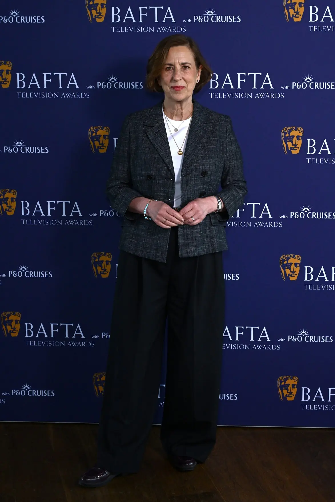 A 70-year-old white woman with a short brunette bob stands in front of a BAFTA branding board. She wears a dark blazer and trousers and a white t shirt.
