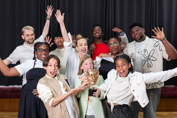 A group of young people, with two adults (Katie Piper and Ben Doyle) stand smiling around a girl holding a BAFTA mask.