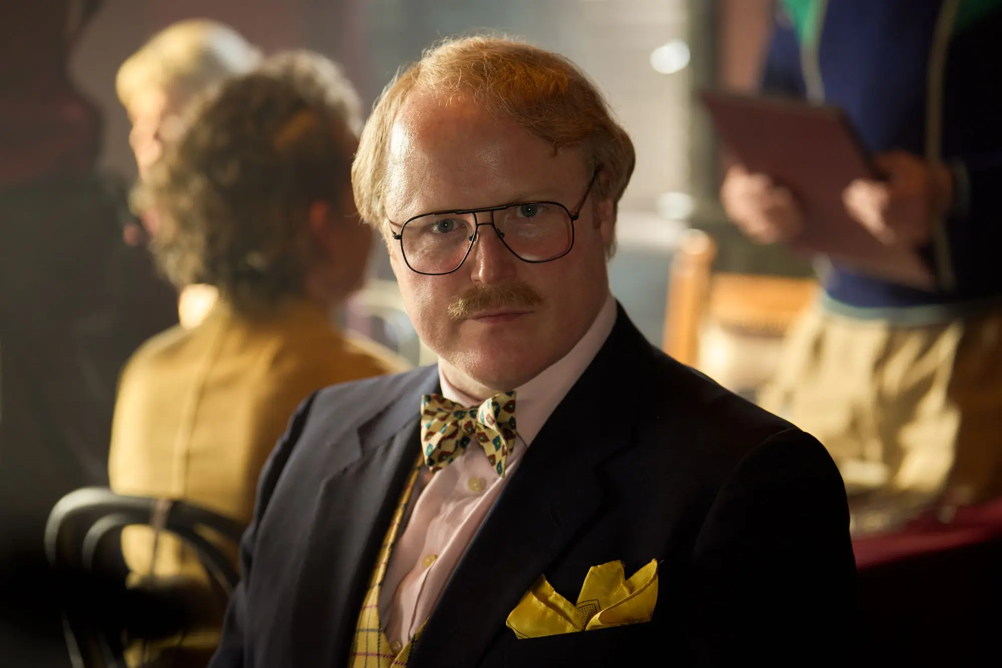 A still of Gary Lamont in Rivals. A man with strawberry blond hair, parted to one side, wearing black-rimmed aviator glasses, a dark blazer and yellow-toned waistcoat, bowtie and pocket square.