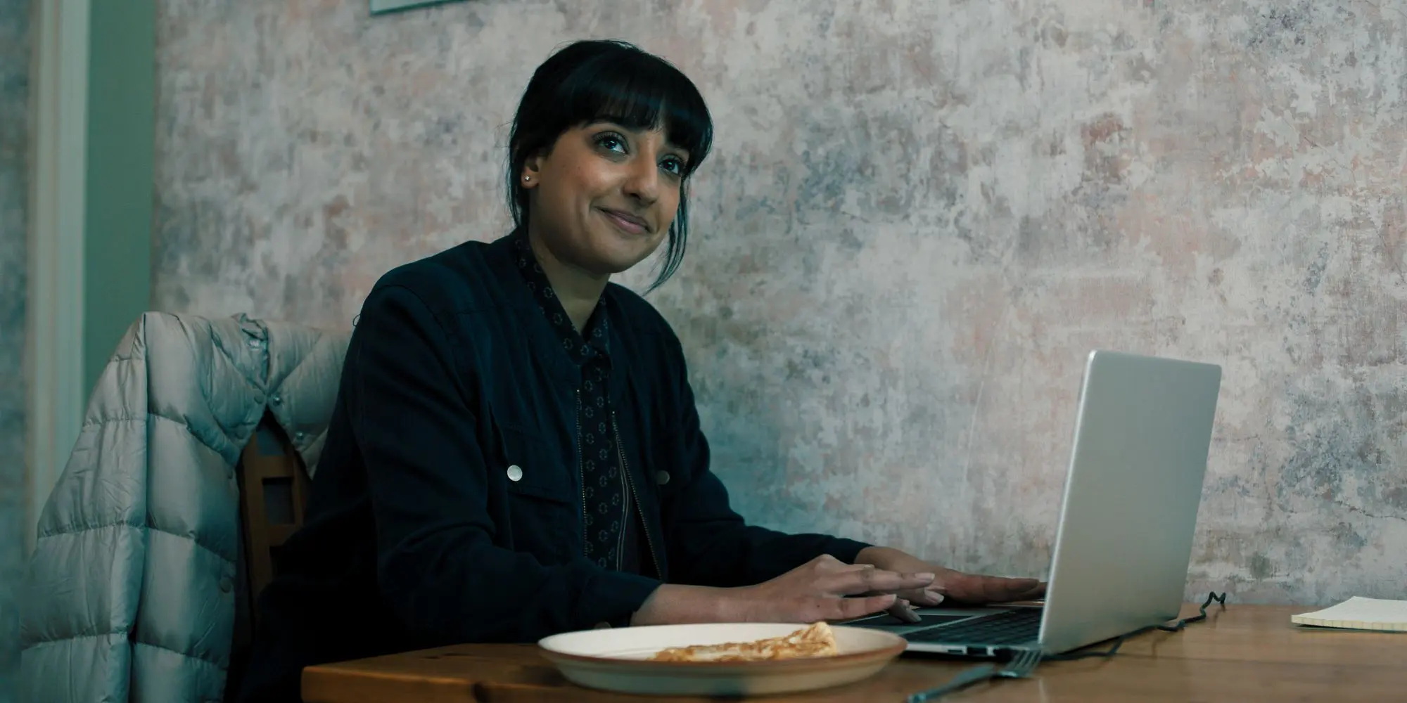 A still of Kiran Sonia Sawar in Slow Horses. An Asian woman with black hair and a thick fringe, wearing all black and sitting in front of a laptop with a plate of food next to her.
