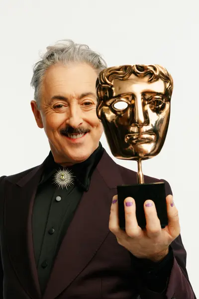 Alan Cumming smiling and holding a BAFTA Award mask next to his face