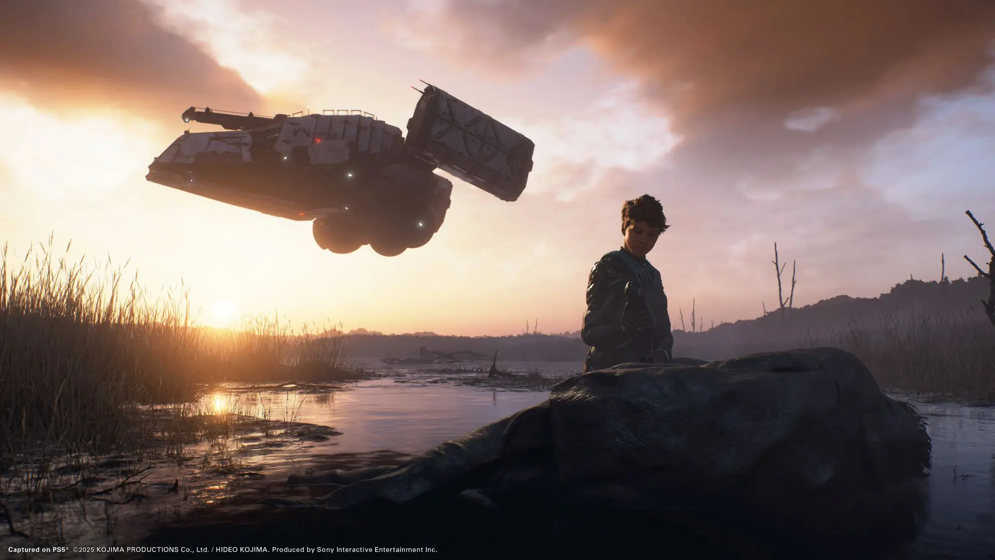 A still from the game Death Stranding 2 On The Beach. A lone figure is stood in what looks like a swamp looking at a fallen washed up figure on the bank. Above is a flying vehicle of some sort.