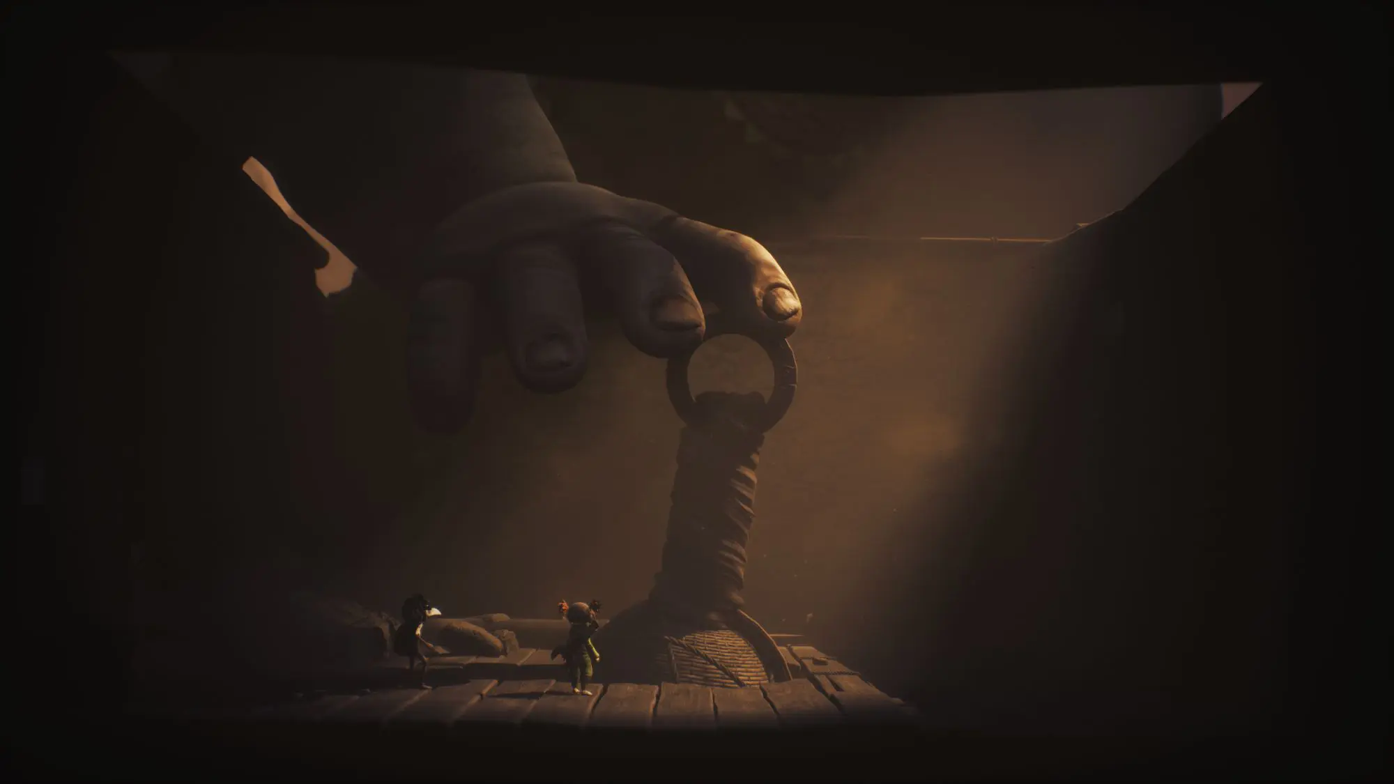 A still from the game Little Nightmares III. Two tiny figures crouch down in what looks like a dark crate as a giant hand reaches in from above.